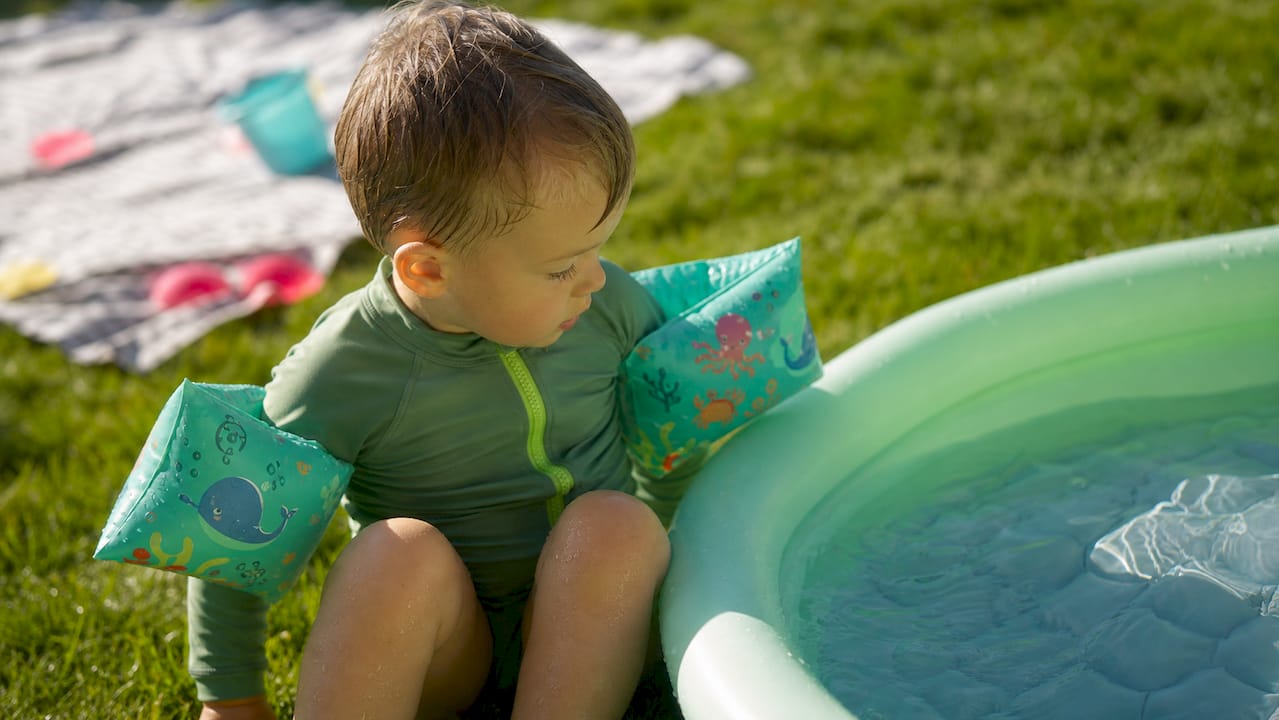 Ein kleines Kind mit Schwimmflügeln sitzt neugierig am Beckenrand und beobachtet das Wasser.